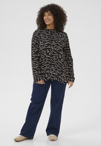 Haut noir à manches longues avec un motif ondulé beige, associé à un jean large bleu foncé et des baskets beiges sur un fond uni.