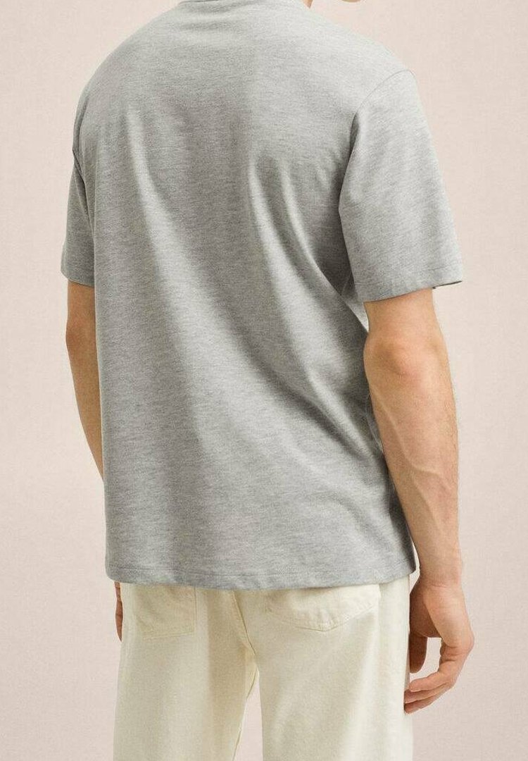 Homme portant une chemise unie gris clair à manches courtes et un pantalon blanc cassé, debout, dos à la caméra.