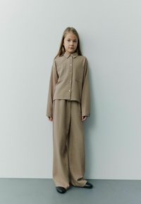 Beige tvådelad outfit bestående av en knapp-up, kortärmad skjorta med två fickor och lösa byxor, tillsammans med svarta skor, mot en ljus bakgrund.