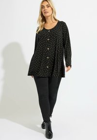 Tunique noire avec des pois dorés, présentant un col rond et une fermeture à boutons. Manches longues et coupe ample avec texture côtelée.