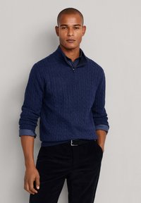 Granatowy sweter w splot kablowy z zamkiem błyskawicznym, noszony na niebieskiej koszuli w kratę. Czarna para spodni z paskiem, gładka struktura i swobodny krój.
