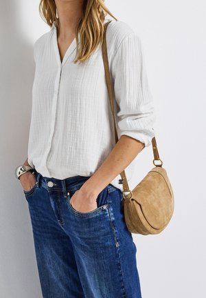 Vrouw die een witte, getextureerde lange mouwen blouse draagt, blauwe jeans, zilveren armband en een beige suède crossbody tas, met handen in de zakken.