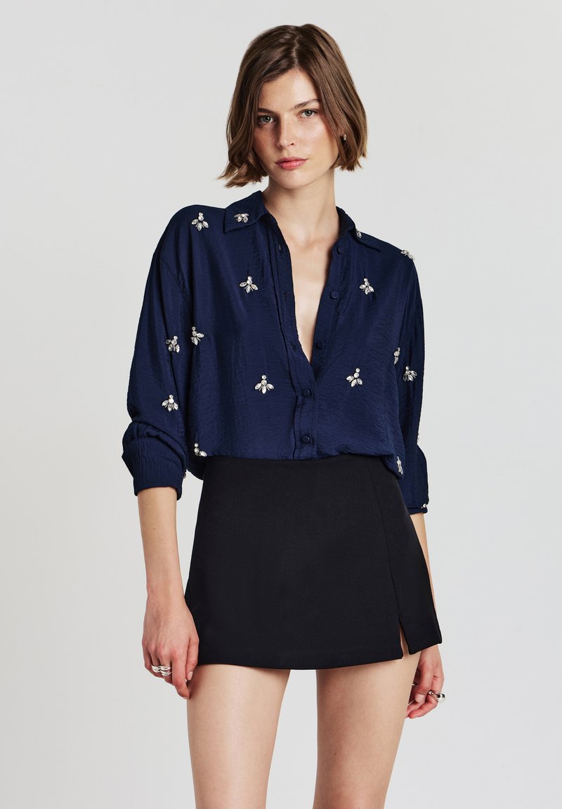 Camicia blu navy con bottoni e motivi di api ricamati in argento, caratterizzata da una vestibilità morbida e maniche lunghe, abbinata a una gonna nera corta.