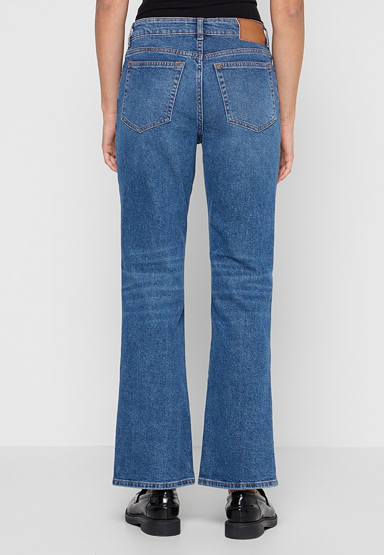 Persoon in mid-blauwe bootcut jeans en zwarte schoenen, van achteren weergegeven tegen een effen lichte achtergrond.