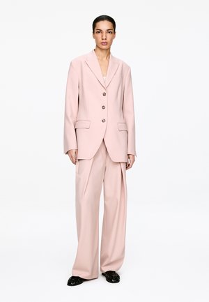 Frau steht frontal, trägt ein hellrosa übergroßes Blazer und passende weit geschnittene Hosen mit schwarzen Schuhen auf weißem Hintergrund.