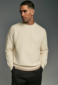 Cremefarvet tekstureret sweatshirt med rund hals og ribbede manchetter, parret med mørke bukser. Simpelt design uden synlige mønstre.