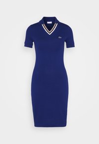 Dunkelblaues Polokleid mit schmaler Silhouette, kurzen Ärmeln und V-Ausschnitt. Mit weißen und beigen Streifenakzenten am Kragen. Lacoste-Logo.