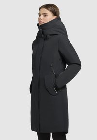 Manteau noir longueur genoux avec un col haut, fermetures éclair et à pression, poches latérales et une texture lisse et mate. Accentué par des zippers.
