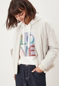 Lichtbeige bomberjack met ribbeldetails; gedragen over een witte hoodie met gekleurde letters; donkere spijkerbroek.