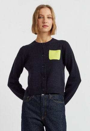 Cardigan a maglia navy con maniche lunghe, colletto tondo, chiusura con bottoni e un dettaglio della tasca di un giallo brillante. Texture liscia e design aderente.