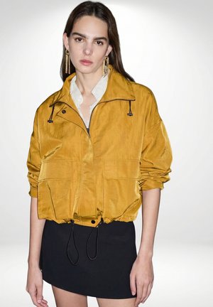 Jonge vrouw met lang donker haar draagt gouden oorbellen, een mosterdgele jas met rits, een wit shirt en een zwarte rok tegen een effen achtergrond.