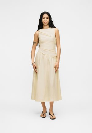 Woman with long black hair wearing a sleeveless beige midi dress and black sandals, standing with relaxed posture on a white background.