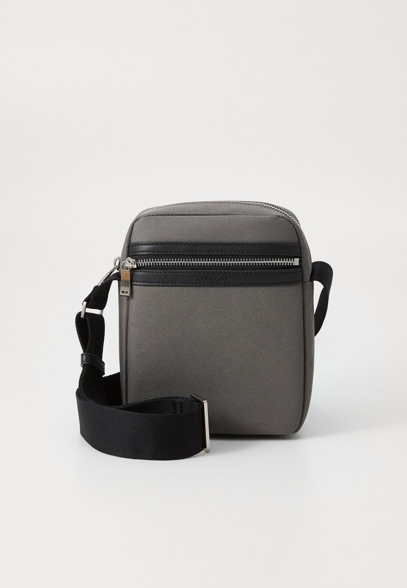 Borsa a tracolla grigia in tessuto con una tasca nera con zip, tracolla regolabile e forma rettangolare. Presenta una texture liscia e accessori in metallo.