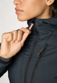 Veste isolante noire avec un col haut, fermeture éclair sur le devant et tirette. Dispose d'une fermeture éclair texturée et d'un accent à cordon.