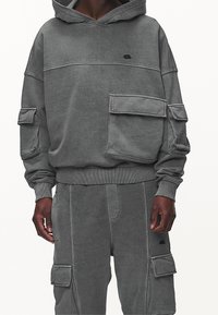 Mörkgrå huvtröja och matchande cargobyxor med stora lockfickor, bärs av en person med mörk hud, visad från axlar till mitten av låret.