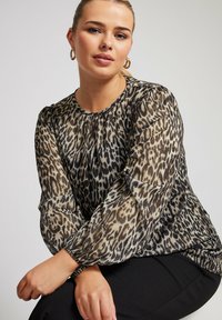 Blusa con stampa leopardata, maniche lunghe e trasparenti; scollatura arricciata; vestibilità comoda; tonalità di marrone e nero; abbinata a pantaloni neri.