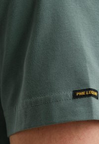 Zelené bavlnené tričko s hladkou textúrou, s čiernou nálepkou s nápisom "PME LEGEND" v žltej farbe na leme. Klasický dizajn s okrúhlym výstrihom.