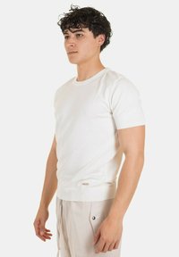 Maglione bianco leggero e aderente a maniche corte con texture a costine e un piccolo logo sul bordo. Materiale morbido e colletto a girocollo classico.