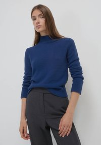 Blauer Pullover mit hohem Kragen, strukturiertem Strickstoff und langen Ärmeln, kombiniert mit grauen, maßgeschneiderten Hosen. Einfaches und sauberes Design.
