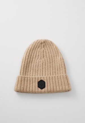 Beige stickad mössa med ribbad struktur, med en vikbar manschett och en svart hexagonal lapp med logotypdetalj.