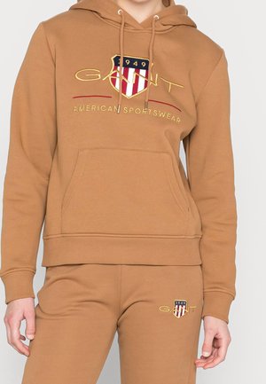 Sweat à capuche marron et pantalon de jogging assorti avec le logo GANT et un écusson présentant des bandes rouges et blanches ainsi que le texte « 1949 ».