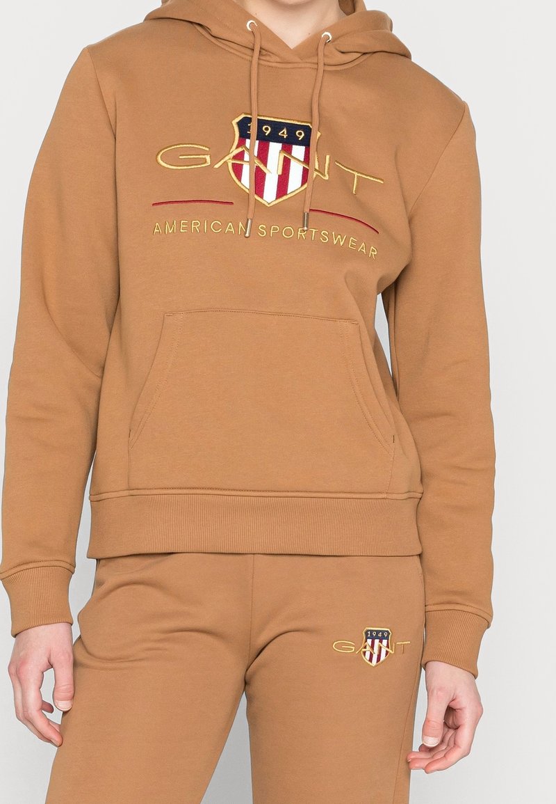 Sweat à capuche marron et pantalon de jogging assorti avec le logo GANT et un écusson présentant des bandes rouges et blanches ainsi que le texte « 1949 ».