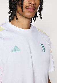 Witte T-shirt met korte mouwen, voorzien van lichtblauwe Adidas- en Juventus-logo's, gele strepen op de schouders en subtiele roze accenten op de naden.