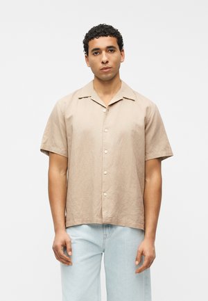 Mies, jolla on lyhyet kiharat hiukset, pukeutunut beigeen lyhythihaiseen napitettuun paitaan ja vaaleansiniset farkut, seisoo valkoista taustaa vasten.