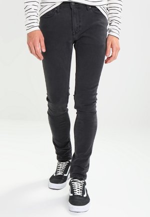 Personne portant un jean noir slim, des baskets noires et blanches, et une chemise blanche à manches longues avec des rayures horizontales noires.