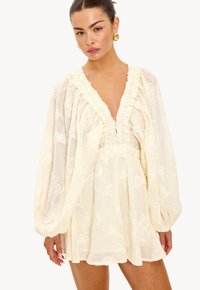 Vestido de crema claro con un escote en V profundo, adornos con volantes, mangas abullonadas y textura floral. Fabricado con una tela suave y ligera.