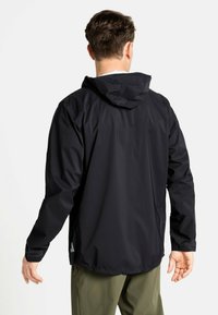 Veste imperméable noire avec capuche, texture lisse, poignets ajustables et logo réfléchissant près de l'ourlet, portée avec un pantalon vert olive.