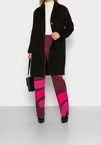 Cappotto lungo nero sopra un dolcevita bianco a coste, abbinato a pantaloni color granata con audaci motivi rosa e stivaletti neri alla caviglia. Inclusa borsa nera.