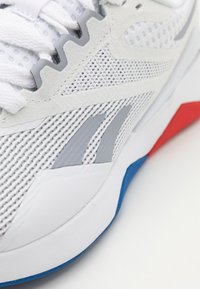 Vitt sportskor med meshtyg på ovansidan, grå och blå detaljer samt röd detalj vid hälen; texturerade ytor och vadderad sula syns.