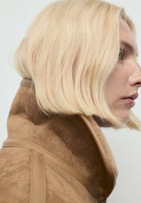 Brązowa zamszowa kurtka z wysokim kołnierzem, z gładkimi beżowymi akcentami. Modelka prezentuje krótkie, falowane blond włosy, zwrócona w lewo.