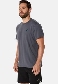 Grå kortærmet t-shirt lavet af bomuldsblandingsstof, med et lille grønt logo på venstre bryst. Matcher med sorte shorts.