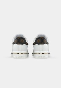 Valged sneakersmustreeritud musta kanna, millel on kuldsed aktsendid tallal ja "GUESS" logo graveeritud taga.