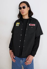 Black short-sleeve shirt with silver snaps, featuring two colorful embroidered patches. Worn over a black long-sleeve top, paired with blue jeans.