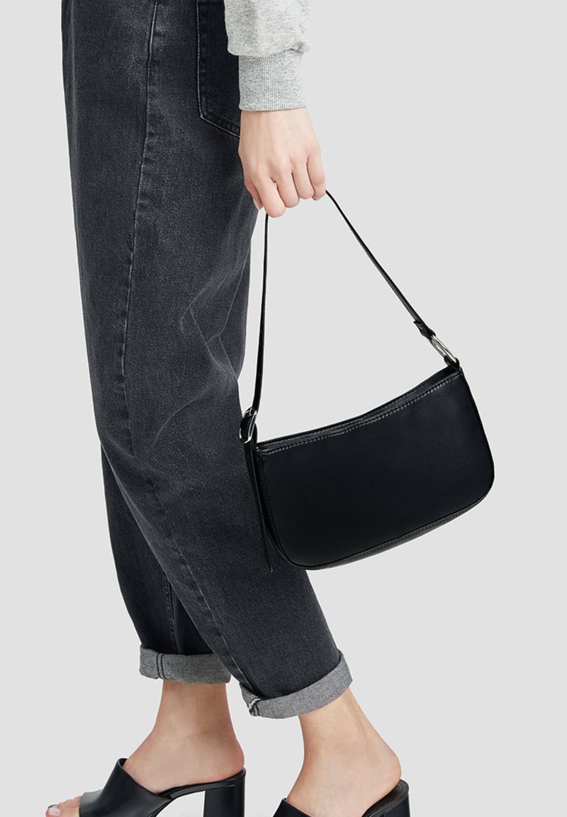 Personne portant un jean noir retroussé et des mules noires à talons, tenant un petit sac à main noir par sa anse sur un fond uni.