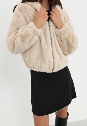 Kvinde iført beige, fluffy lynlåstrøje med hætte over et sort skørt og sorte støvler, som holder jakken tæt ved kraven.