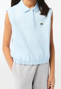 Světle modré dámské polo bez rukávů s žebrovaným límečkem, dvěma knoflíky ve výstřihu a zeleným logem na prsou. Měkká, strukturovaná látka se shromažďuje u spodního lemu.