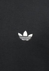 Λευκό λογότυπο Adidas trefoil κεντημένο σε μαύρο πλεκτό φόντο, κεντραρισμένο και ευδιάκριτο.