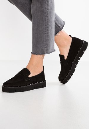 Czarne zamszowe buty wsuwane z grubą czarną podeszwą i srebrnymi ćwiekami wzdłuż krawędzi, zestawione z szarymi spodniami o skróconej długości.