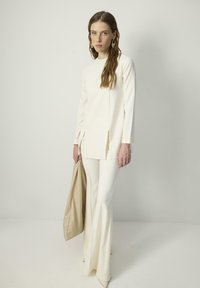 Cream-colored long-sleeve top with side slits, paired with flared cream pants. Holding a beige bag. Flat pointed-toe shoes. Smooth texture.