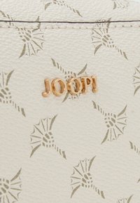 Matériau blanc texturé présentant un motif répétitif de fleurs stylisées. Logo doré "JOOP!" affiché de manière proéminente.