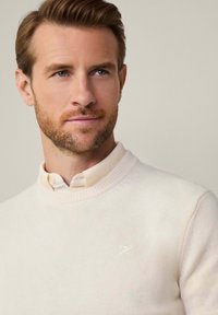 Svetr v světlé krémové barvě, vyrobený z měkkého pleteného materiálu. Má kulatý výstřih a diskrétní logo na levé části hrudi. Texturovaný povrch.