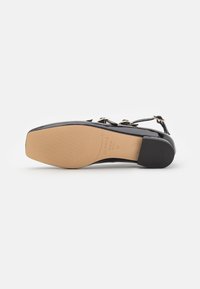 Svarte patentlær flate sandaler med spiss tå, gull hardware-detaljer og gummihæl. Sålen er beige med et teksturert gripe mønster.