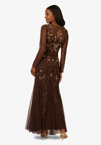 Hnědá večerní šaty s dlouhými rukávy a přiléhavým korzetem, tylovou sukní a složitou zlatou květinovou výšivkou. Design doplňuje detail s klíčovou dírkou na zádech.