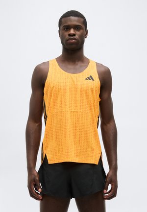 Muž stojaci v oranžovom športovom tričku bez rukávov a čiernych šortkách na jednoduchom svetlom pozadí.