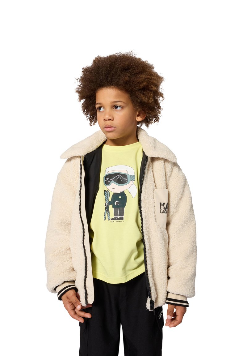 Enfant aux cheveux bouclés portant une veste polaire beige par-dessus un t-shirt jaune avec un motif graphique et un pantalon noir, regardant légèrement sur le côté.