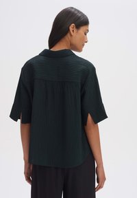 Dark green shirt with a textured, ribbed design. Features a collar, short sleeves, and side slits. Loose fit with a slightly curved hem.